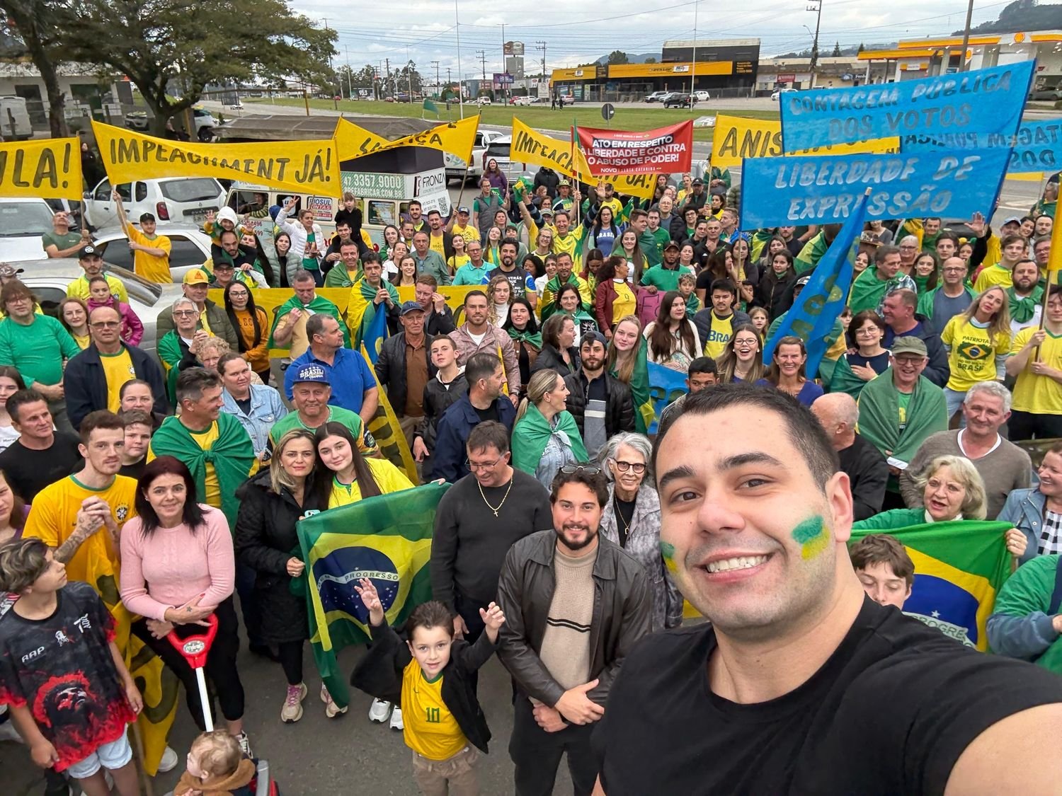 Ato público com centenas de pessoas, bandeiras do Brasil e faixas, com Lucas Jean em primeiro plano.