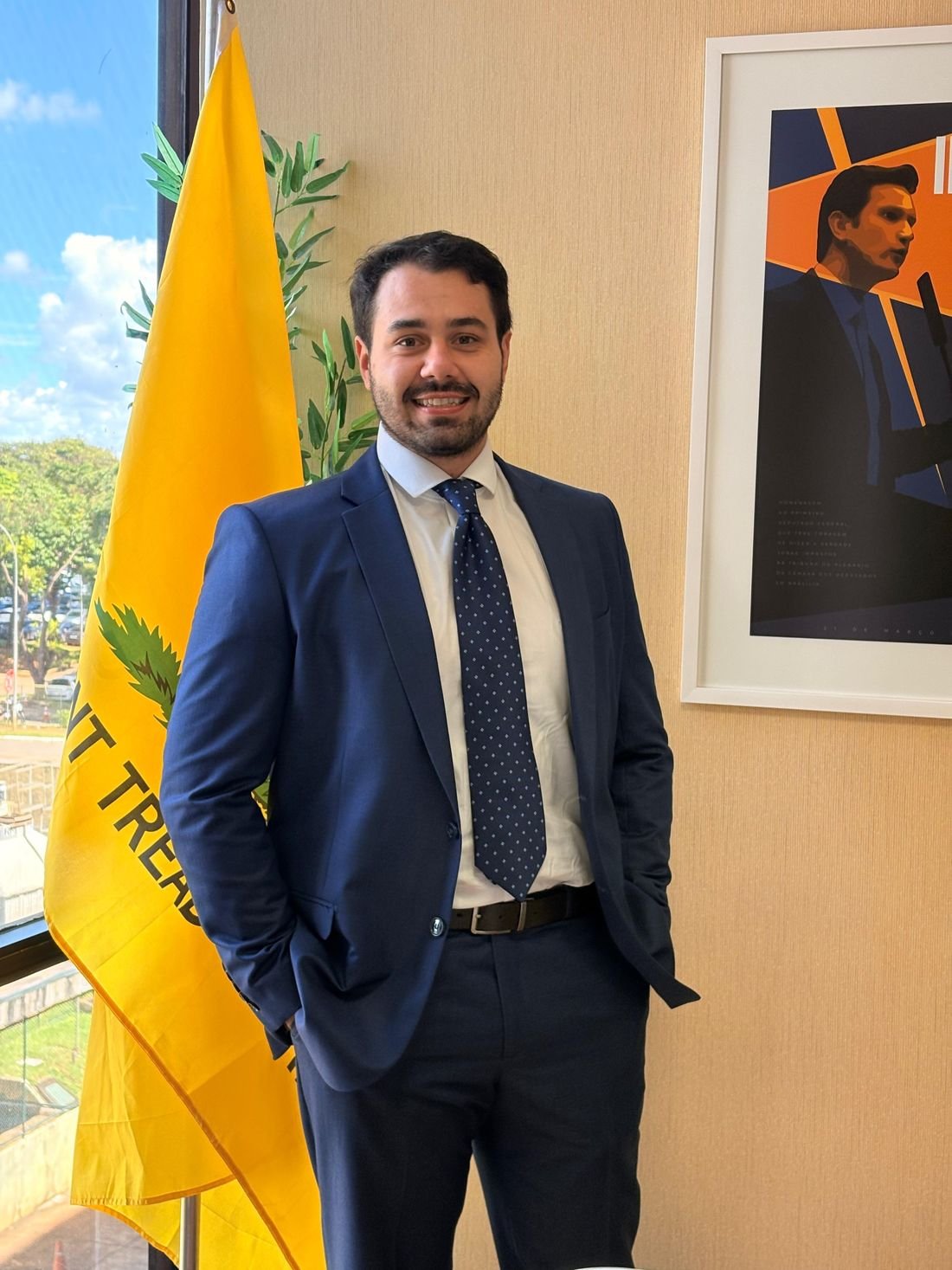 Lucas Jean de terno, em pé ao lado de uma bandeira, em um gabinete.