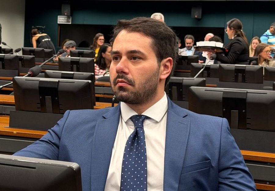 Lucas Jean sentado em uma sessão, em plenário com bancadas e microfones.
