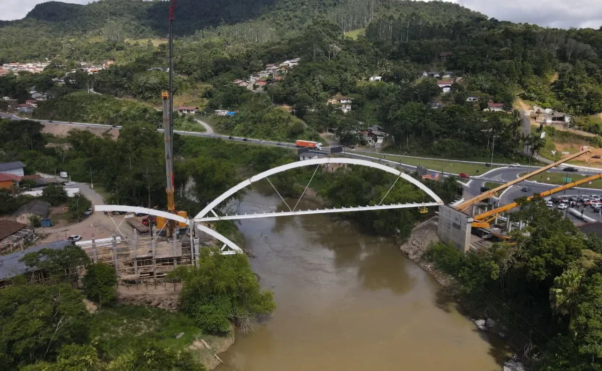 Vista aérea da ponte em arco em obras, sobre o rio, com guindastes nas duas margens.